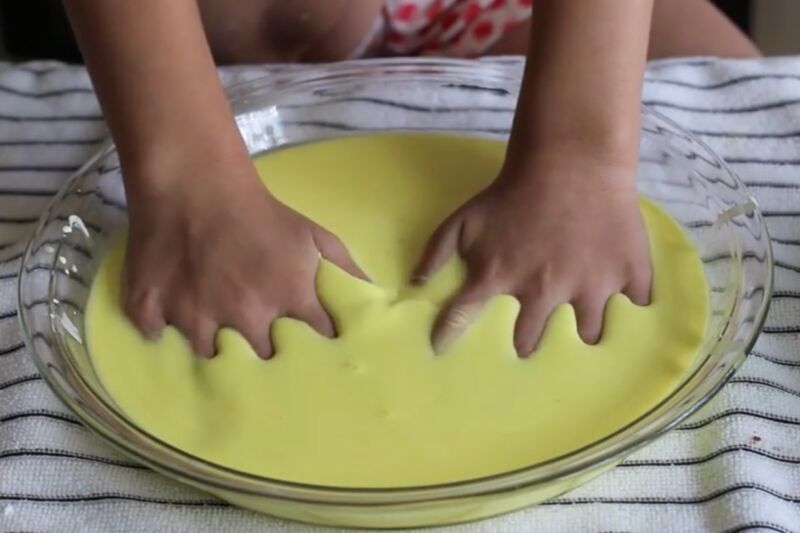 child's hands pressing into a yellow gooey substance in a glass bowl.